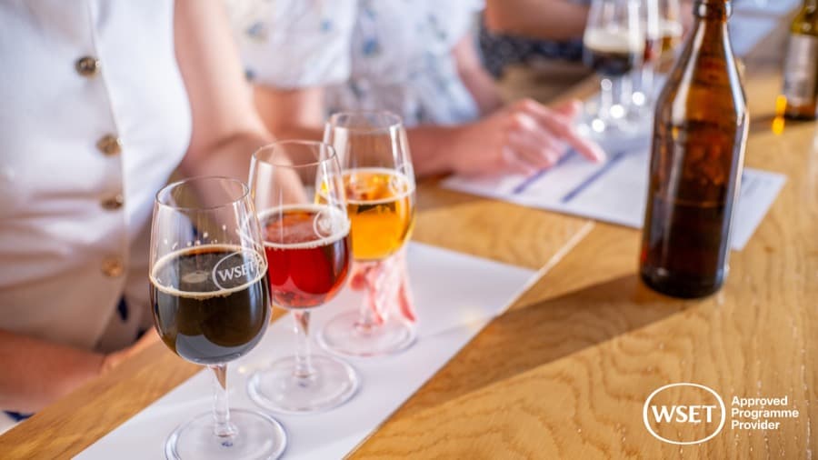 Três cervejas de cores diferentes em taças de degustação, ao lado de uma garrafa, sobre uma mesa de madeira