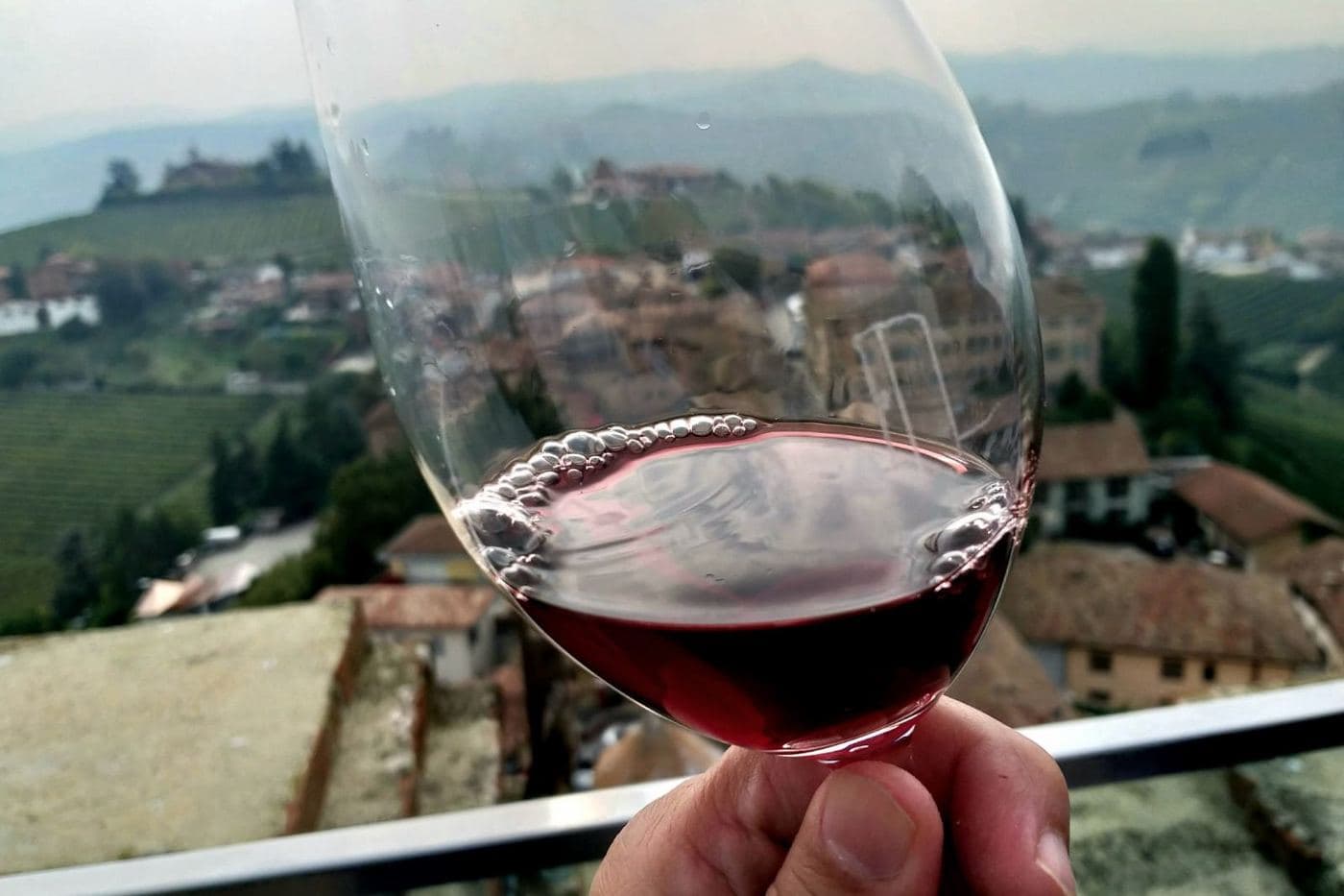 Taça de vinho tinto diante de vilarejo e vinhedos das Langhe